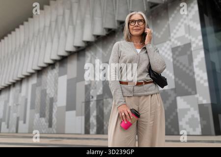 Woman in casual attire stands outdoors with coffee while using a smartphone in a modern urban setting Stock Photo
