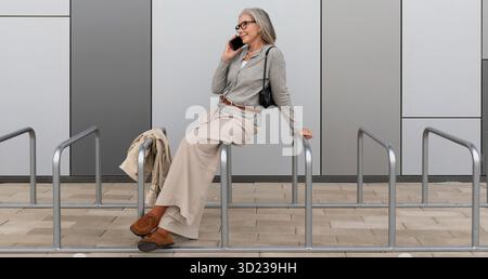 Older woman casually talking on mobile phone while sitting on railings outside modern building Stock Photo
