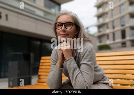 Senior woman enjoys a moment of reflection on a bench in a modern urban setting during a cloudy afternoon Stock Photo