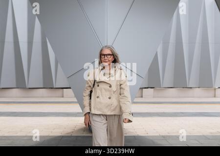 Woman walks confidently in stylish trench coat near modern architectural structure during daylight Stock Photo