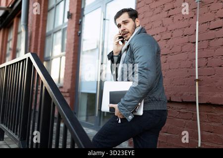 A male programmer talking on the phone with a laptop in his hands enters the office building Stock Photo