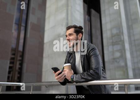 Young stylish man in a leather jacket with a cup of coffee by a mobile phone on the background of a gray city building Stock Photo