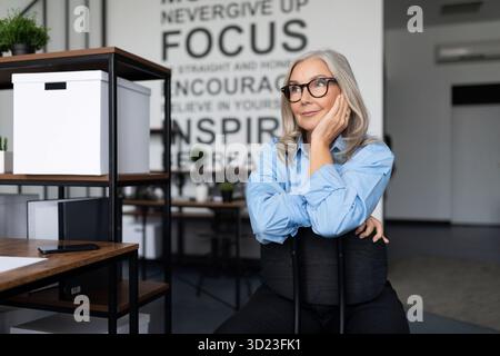 Caucasian senior businesswoman in a blue shirt and glasses sits thinking in the office Stock Photo