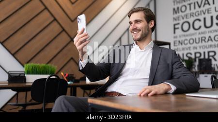Young businessman sitting at a table in the office talking on a video call using a mobile phone Stock Photo
