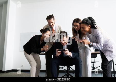 A team of professional office workers stands around a colleague playing on a mobile phone in the office Stock Photo