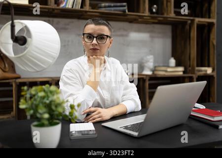 Caucasian stylish adult woman with short haircut in glasses sitting at laptop in stylish office Stock Photo