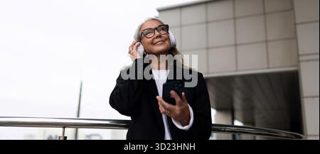Elderly stylish woman listens to music in headphones with a phone in her hands against the backdrop of the building Stock Photo
