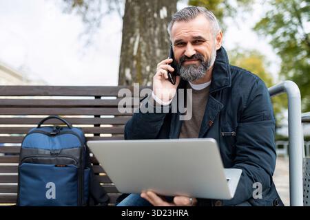 An elderly business man speaks on a mobile phone while working on a laptop online in a park on a bench Stock Photo