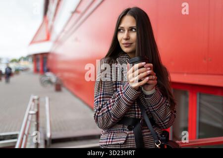 A young woman with a cup of tea in her hands warms her hands with a smile looking into the distance next to the shopping center Stock Photo