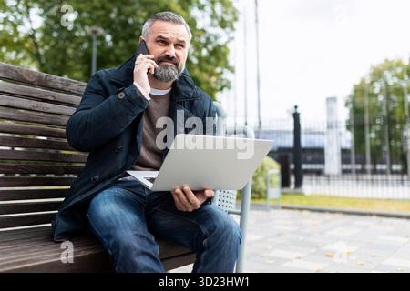 Mature man talking on a mobile phone with a laptop in his hands outside Stock Photo