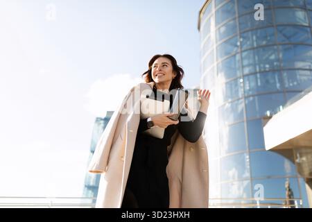 Portrait of a successful young business woman in a stylish coat against the backdrop of an office skyscraper, woman leader in bu Stock Photo