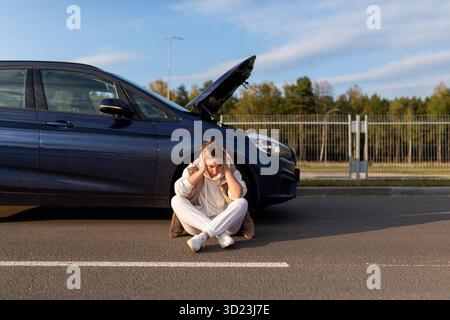 Frustrated woman sits holding her head on the road next to a collapsed car with the hood up Stock Photo