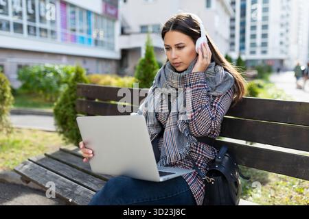 A young woman works online on a laptop and listens to music on headphones while sitting on a bench next to a shopping center Stock Photo