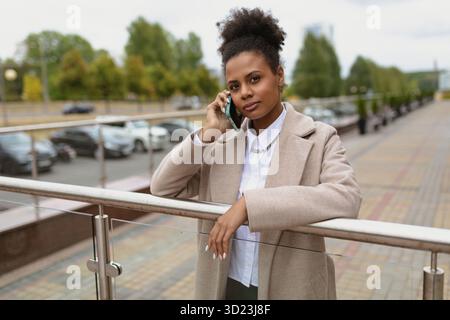 Serious african american woman lawyer in a beige coat on the background of the city speaks on a mobile phone Stock Photo