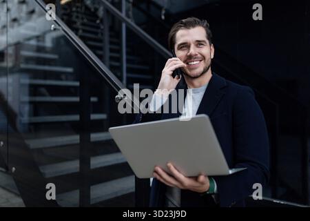 Middle-aged business man speaks on a mobile phone with a wide smile and holds a laptop in his hands, remote work concept Stock Photo