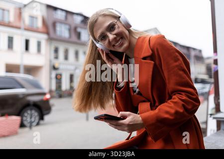 Cheerful female student listens to music in headphones using phone and looks at camera with a smile Stock Photo