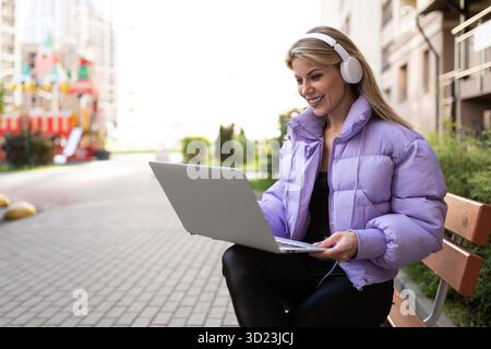 Stylish modern woman working on laptop with headphones outside Stock Photo