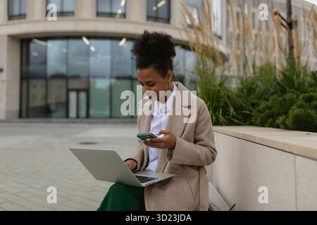 Young african american woman student working online on laptops with mobile phone on the background of urban modern building Stock Photo