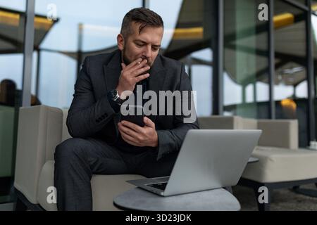 Male businessman working on a laptop online with a mobile phone in his hands sitting outside the office with a cigarette Stock Photo