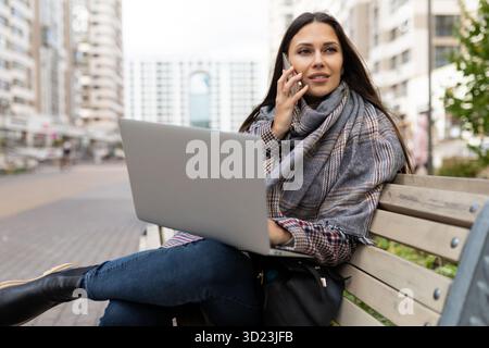 Female freelancer speaks on a mobile phone and works on a laptop Sitting on a bench against the backdrop of city buildings Stock Photo