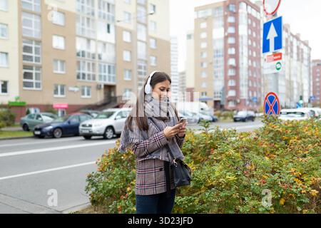 Stylish young woman on a crosswalk in headphones looks at a mobile phone Stock Photo