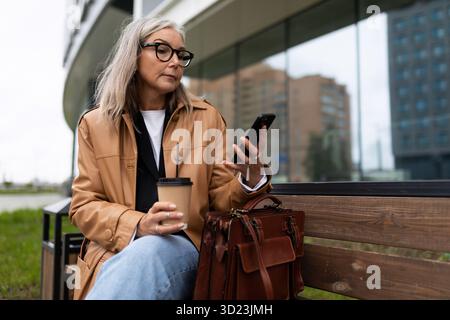 Stylish woman with a cup of coffee in her hands of European appearance looks at a mobile phone outside Stock Photo