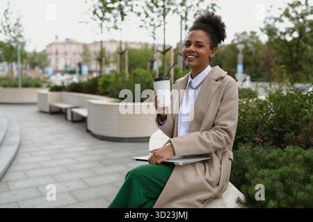 Portrait african american smiling woman with laptop and cup of coffee outside office, concept of successful business development Stock Photo