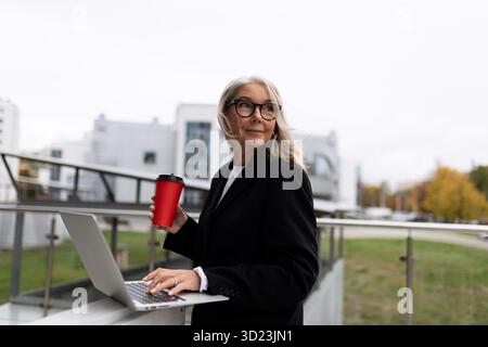 Middle aged older business woman works on a laptop and drinks coffee on the office terrace, concept of successful business devel Stock Photo