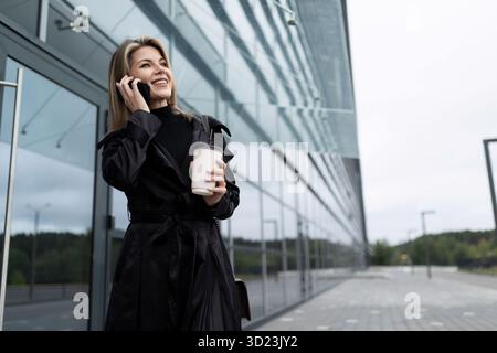 Woman businessman with a cup of coffee on the background of an office glass building talking on a mobile phone with a smile on h Stock Photo