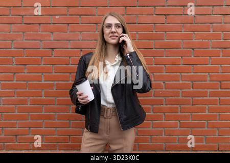 Young female student talking on a mobile phone with a cup of coffee in her hands against the background of an orange brick wall Stock Photo