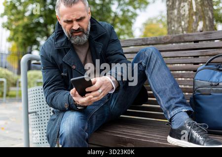 Adult man sitting on a bench in the park looking at a mobile phone Stock Photo