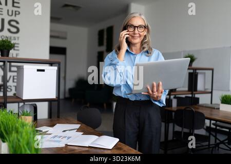Middle aged older business woman in the office talking on a mobile phone with a laptop in her hands Stock Photo