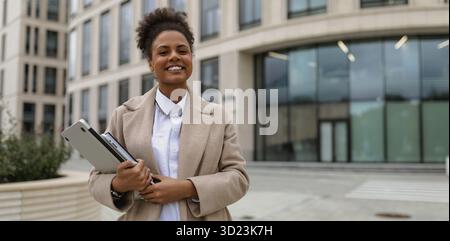 Portrait of a successful young african american woman with a laptop in her hands and a wide smile against the backdrop of an off Stock Photo