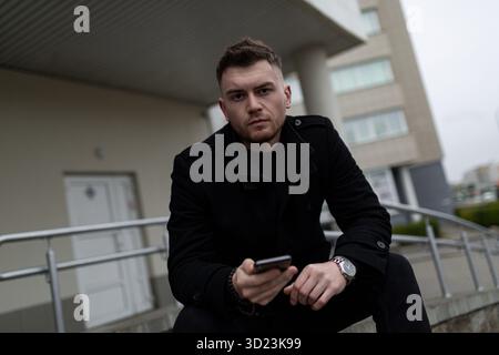 Young man with a phone in his hands sitting looking at the camera outside Stock Photo