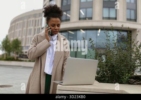 African american business woman talking on a mobile phone and answering an email on a laptop online against the backdrop of a ci Stock Photo