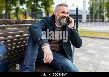 A mature man with a gray beard is talking on the phone with a smile while sitting on a bench in a city park Stock Photo
