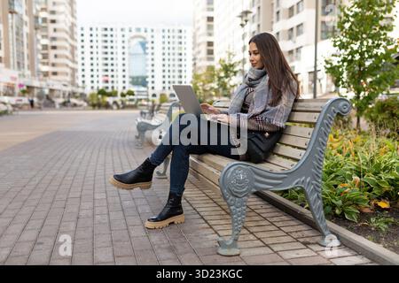 Young woman working on a laptop online sitting in a city park against the backdrop of residential buildings, Concept freelancing Stock Photo