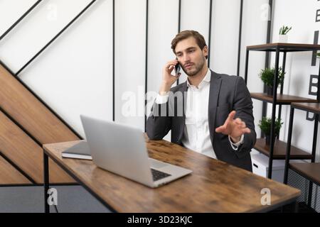 Young businessman sitting at a laptop in the office talking on a mobile phone Stock Photo