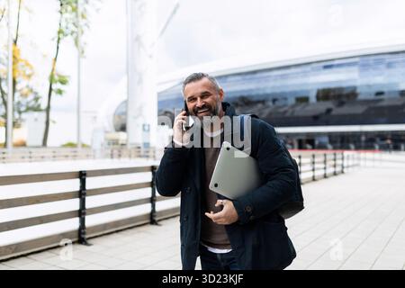 A man with a laptop in his hands talking on the phone walking down the street Stock Photo