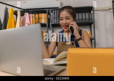 Young female entrepreneur smiling while talking on the phone and using a laptop at home. She manages an online store, checks orders, and communicates Stock Photo