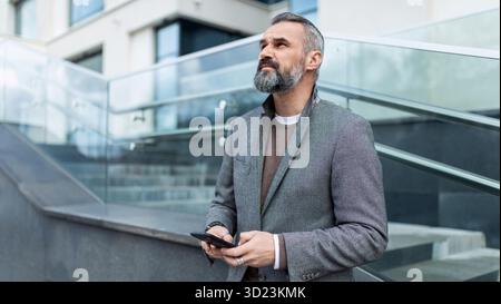 Adult confident businessman With a slight smile on his face looks up with a mobile phone in his hands on the background of a bus Stock Photo
