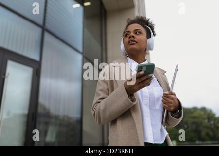 Stylish african american female student in headphones using mobile phone and laptop on the background of the glass facade of the Stock Photo