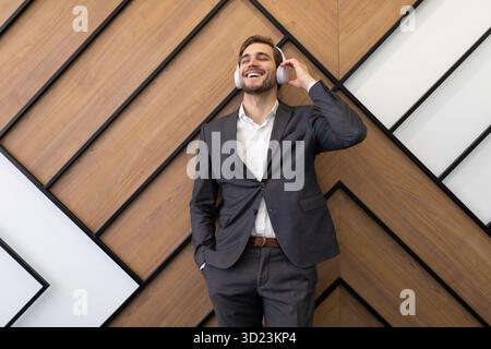 A young businessman in a strict gray suit enjoys listening to music on headphones against the background of an office wall Stock Photo