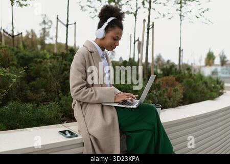 Young black female student preparing for an exam working on a laptop online in headphones in a city park Stock Photo
