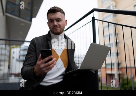 Young businessman entrepreneur working on a laptop online outside the office with a mobile phone in his hands Stock Photo