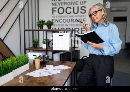 Confident woman accountant speaks on a mobile phone with a notepad for writing in her hands against the backdrop of a trendy off Stock Photo