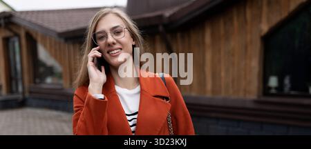 Stylish young businesswoman speaks on a mobile phone in a bright orange coat on the background of a modern building Stock Photo