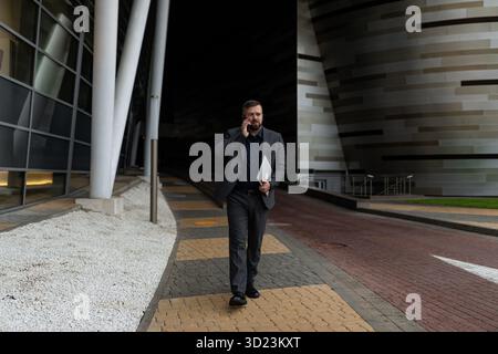 Mature business man in a business suit talking on the phone with a laptop in his hands walking along the building Stock Photo