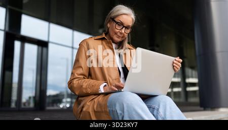 Business senior woman with laptop working online on steps of office business center Stock Photo