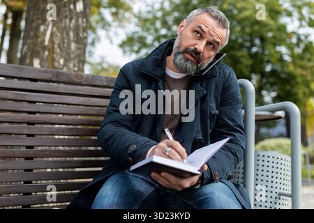 Mature adult man talking on the phone making notes in a diary while sitting in the park Stock Photo